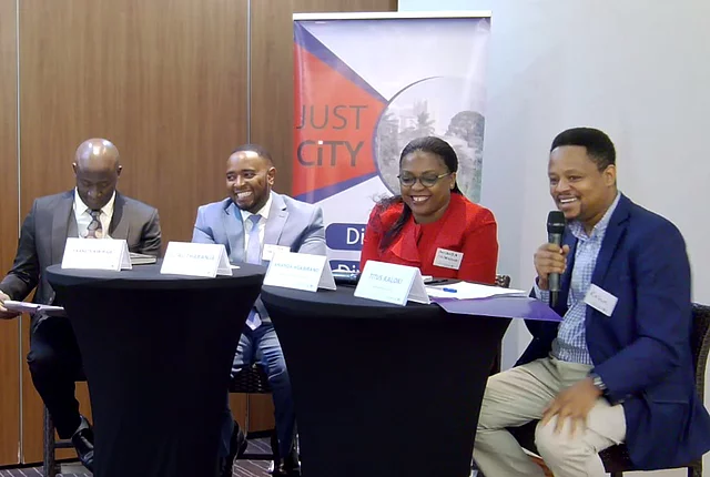 Four individuals seated at two black round tables during a formal panel discussion. Each has a name placard: "ISAAC KURIGIA," "GITHAU THABANJA," "AMANDA NGABIRANO," and "TITUS KALOKI," with the latter speaking into a microphone. Behind them is a banner with the text "JUST CITY" and partially visible wording, suggesting a conference or seminar focused on urban issues.