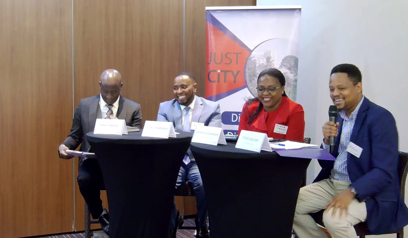 Four individuals seated at two black round tables during a formal panel discussion. Each has a name placard: "ISAAC KURIGIA," "GITHAU THABANJA," "AMANDA NGABIRANO," and "TITUS KALOKI," with the latter speaking into a microphone. Behind them is a banner with the text "JUST CITY" and partially visible wording, suggesting a conference or seminar focused on urban issues.