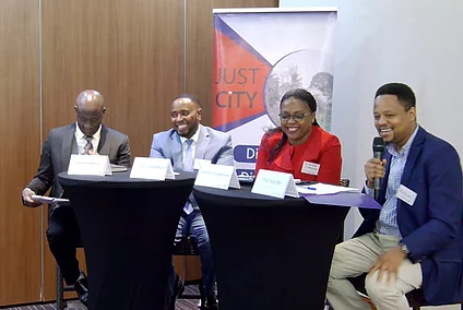 Four individuals seated at two black round tables during a formal panel discussion. Each has a name placard: "ISAAC KURIGIA," "GITHAU THABANJA," "AMANDA NGABIRANO," and "TITUS KALOKI," with the latter speaking into a microphone. Behind them is a banner with the text "JUST CITY" and partially visible wording, suggesting a conference or seminar focused on urban issues.