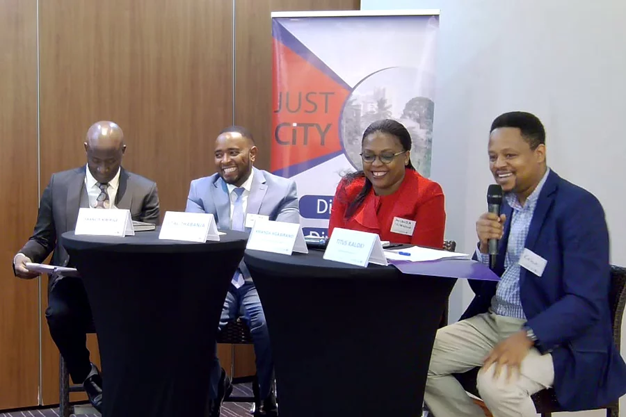 Four individuals seated at two black round tables during a formal panel discussion. Each has a name placard: "ISAAC KURIGIA," "GITHAU THABANJA," "AMANDA NGABIRANO," and "TITUS KALOKI," with the latter speaking into a microphone. Behind them is a banner with the text "JUST CITY" and partially visible wording, suggesting a conference or seminar focused on urban issues.