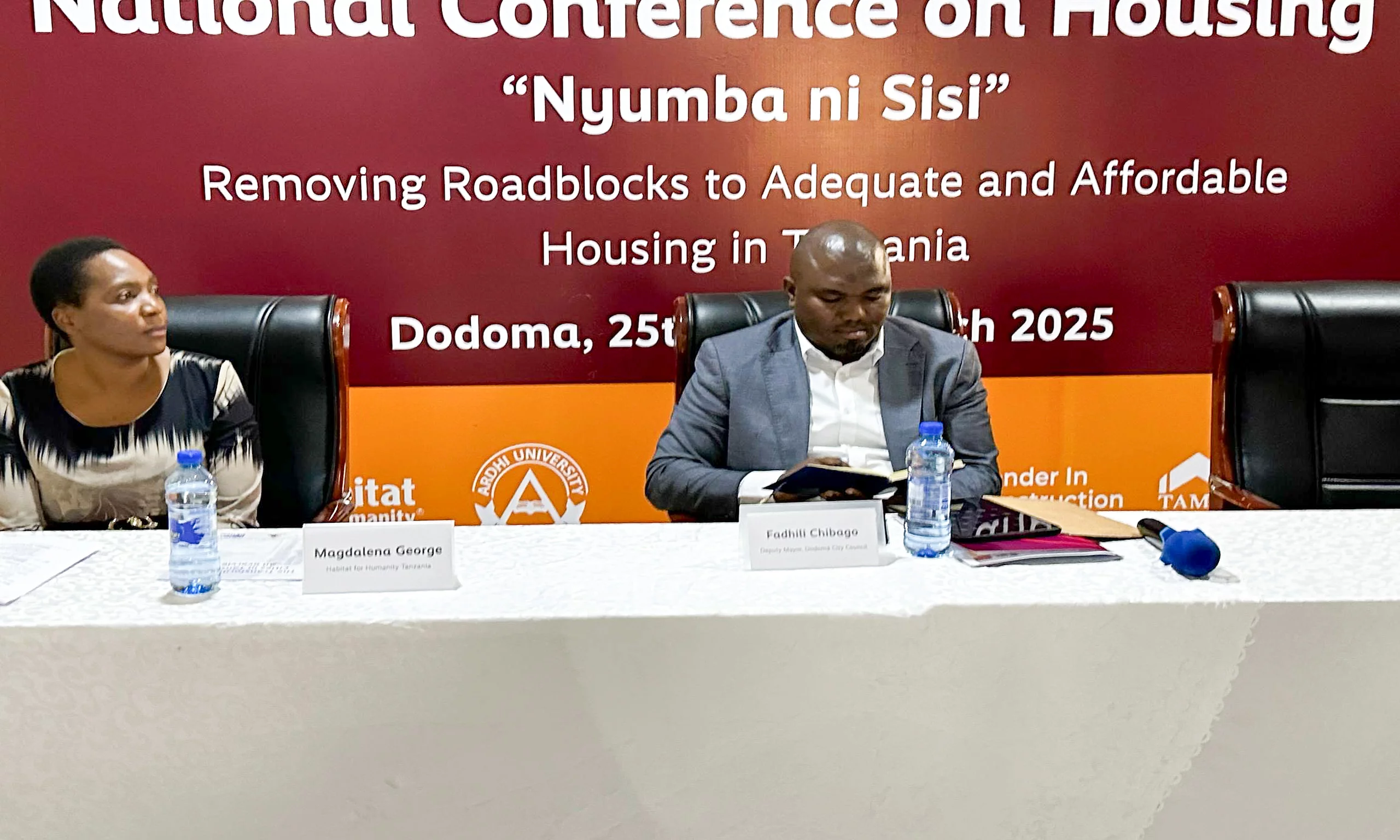 Two individuals seated at a conference table during the "National Conference on Housing" in Dodoma, held on May 24–25, 2023. The backdrop displays the event's theme: “Nyumba ni sisi – Ensuring Decent and Adequate Affordable Housing for All.” Microphones, bottled water, and conference materials suggest a formal setting focused on housing policy and development.