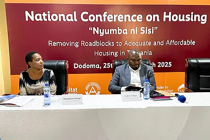 Two individuals seated at a conference table during the "National Conference on Housing" in Dodoma, held on May 24–25, 2023. The backdrop displays the event's theme: “Nyumba ni sisi – Ensuring Decent and Adequate Affordable Housing for All.” Microphones, bottled water, and conference materials suggest a formal setting focused on housing policy and development.