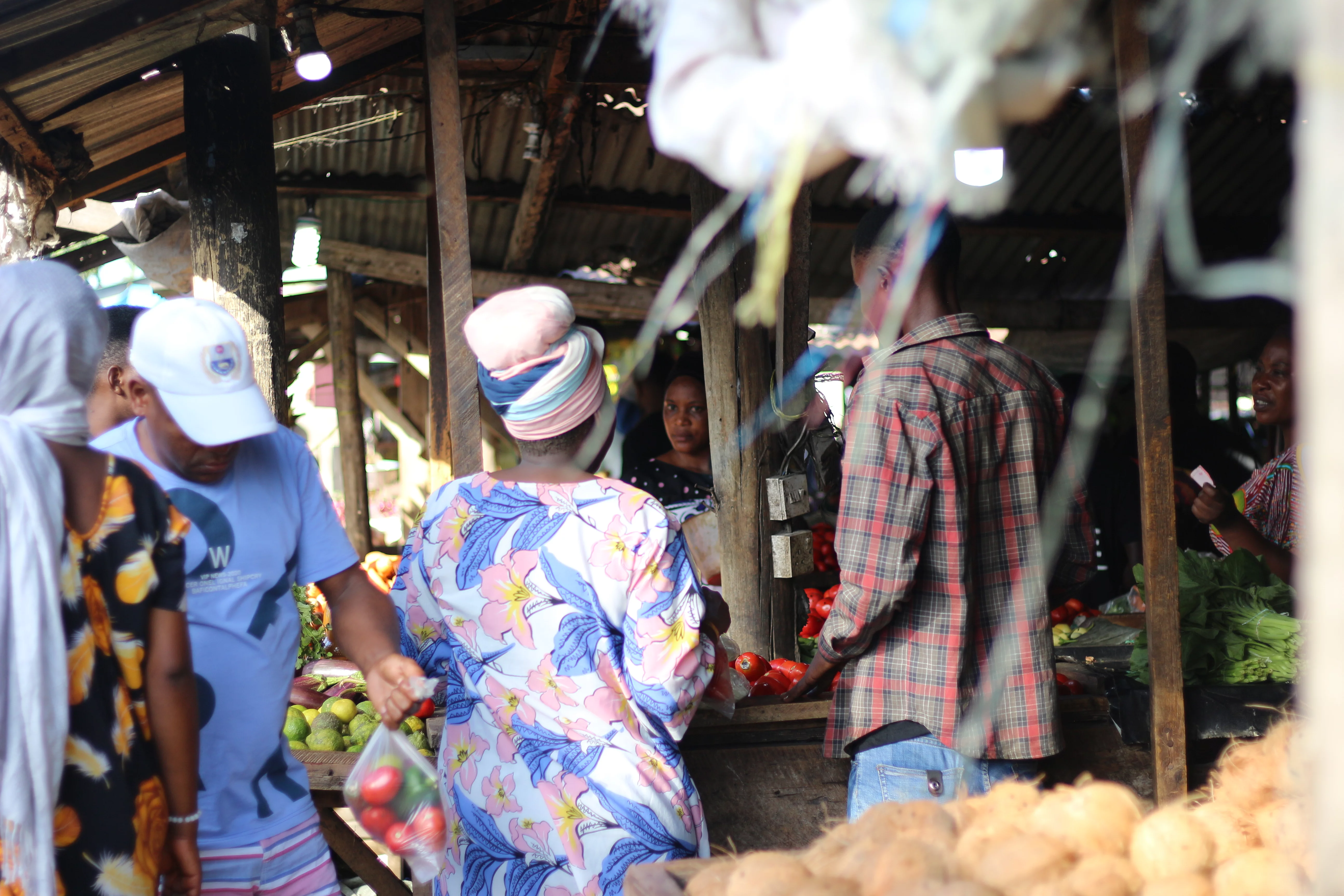 A lively open-air market scene with people shopping and selling under a wooden roofed structure. Individuals wear colorful clothing, and one person holds a transparent bag of produce. Various fruits and vegetables, including a pile of yams or potatoes, are visible in the foreground. The atmosphere reflects vibrant community interaction and daily commerce.
