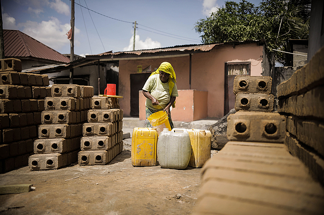 A person wearing a yellow headscarf and green shirt pours water from a yellow bucket into large plastic containers, mostly yellow with one white. The scene takes place outdoors in a residential area with a pink and brown house in the background. Neatly stacked bricks with circular holes and a red plastic cup are visible nearby, set on a dusty ground with utility poles and wires overhead.