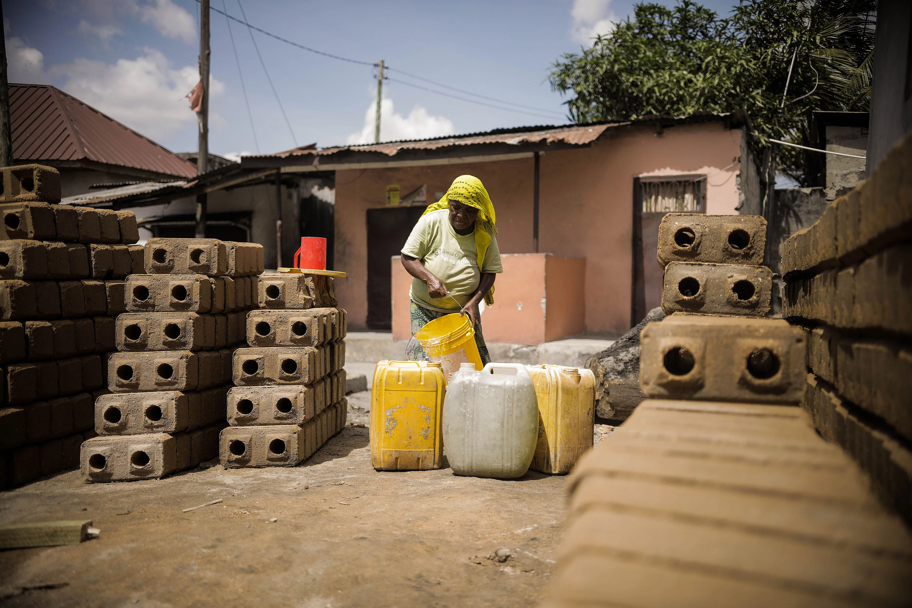 A person wearing a yellow headscarf and green shirt pours water from a yellow bucket into large plastic containers, mostly yellow with one white. The scene takes place outdoors in a residential area with a pink and brown house in the background. Neatly stacked bricks with circular holes and a red plastic cup are visible nearby, set on a dusty ground with utility poles and wires overhead.