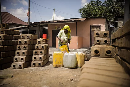 A person wearing a yellow headscarf and green shirt pours water from a yellow bucket into large plastic containers, mostly yellow with one white. The scene takes place outdoors in a residential area with a pink and brown house in the background. Neatly stacked bricks with circular holes and a red plastic cup are visible nearby, set on a dusty ground with utility poles and wires overhead.