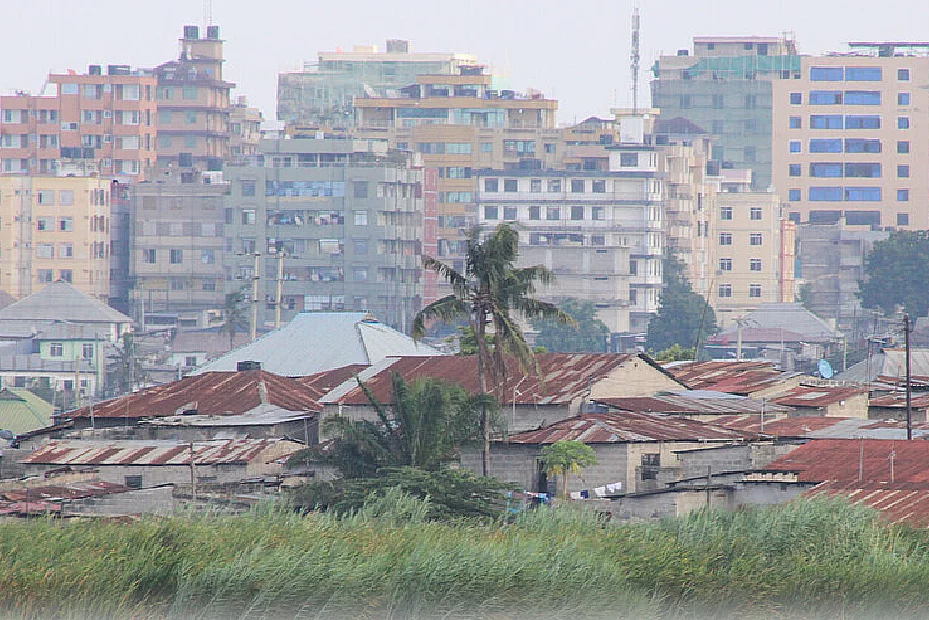 A cityscape showing a contrast between dense green vegetation in the foreground and a mix of low-rise buildings with corrugated metal roofs behind it. In the background, modern multi-story buildings rise, highlighting the socio-economic diversity and urban development within the area.