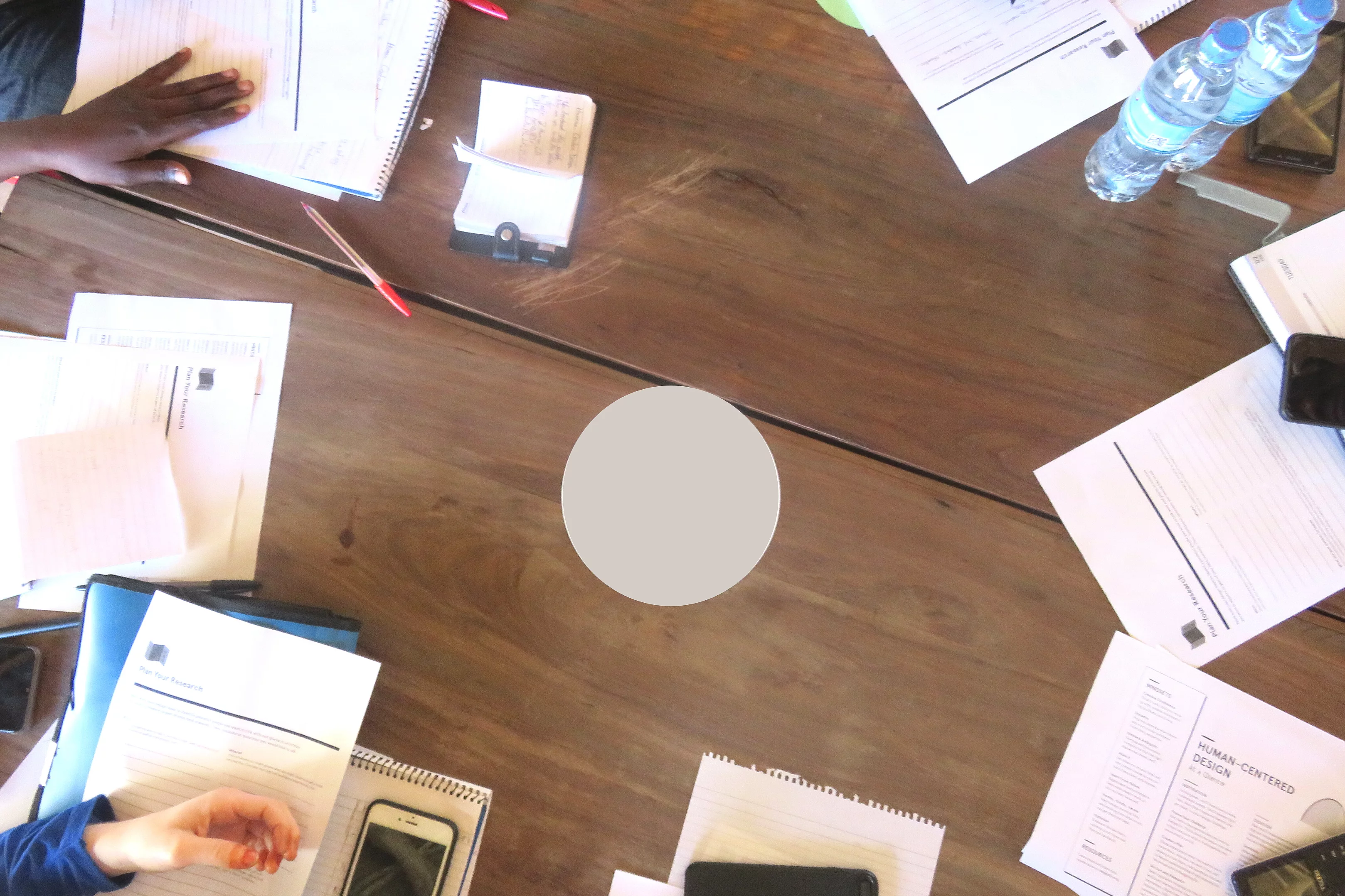 A top-down view of a group of people seated around a wooden table, actively engaged in a collaborative session. Papers with printed text and diagrams, notebooks, pens, mobile phones, and a plastic water bottle are spread across the table. The scene suggests a focused learning or working environment, possibly a workshop or study group.