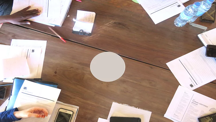 A top-down view of a group of people seated around a wooden table, actively engaged in a collaborative session. Papers with printed text and diagrams, notebooks, pens, mobile phones, and a plastic water bottle are spread across the table. The scene suggests a focused learning or working environment, possibly a workshop or study group.