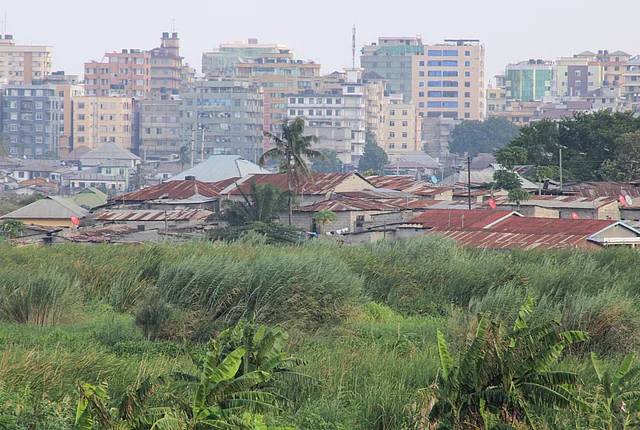 A cityscape showing a contrast between dense green vegetation in the foreground and a mix of low-rise buildings with corrugated metal roofs behind it. In the background, modern multi-story buildings rise, highlighting the socio-economic diversity and urban development within the area.