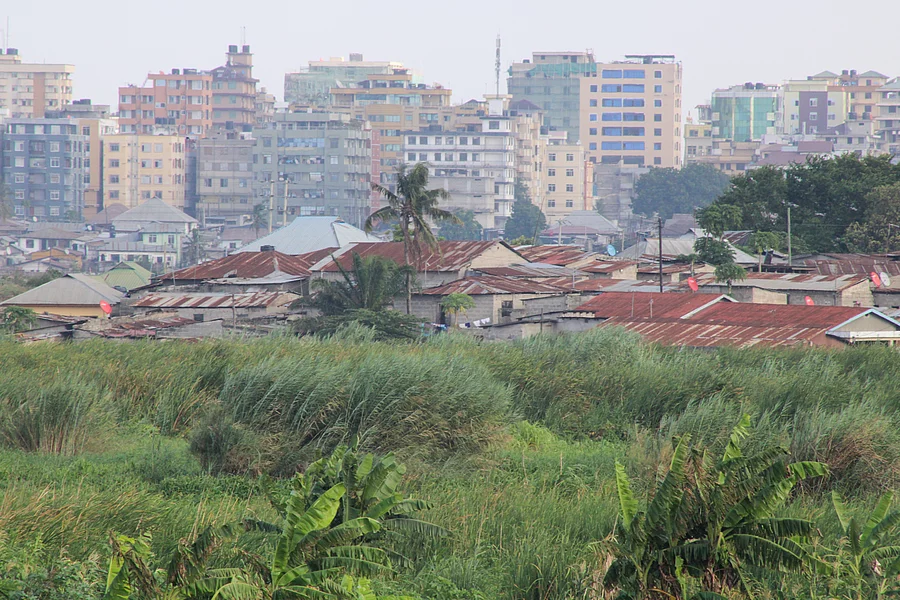 A cityscape showing a contrast between dense green vegetation in the foreground and a mix of low-rise buildings with corrugated metal roofs behind it. In the background, modern multi-story buildings rise, highlighting the socio-economic diversity and urban development within the area.