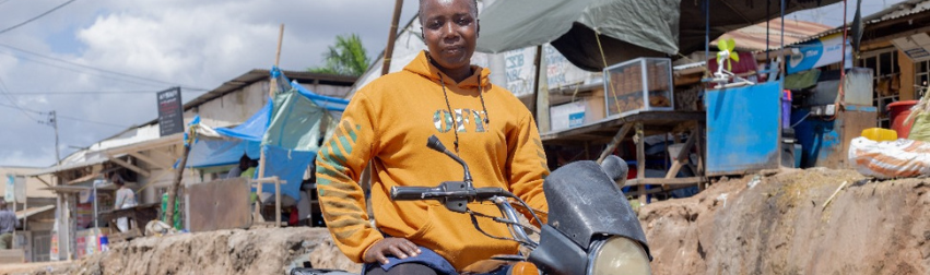 A person wearing a bright orange hoodie with the word "OFF" sits on a black motorcycle parked on a dirt road in a bustling outdoor market. Behind them, wooden and metal stalls covered with tarps display various goods, creating a vibrant and textured backdrop of local commerce. The scene captures everyday life and transportation in a community setting.