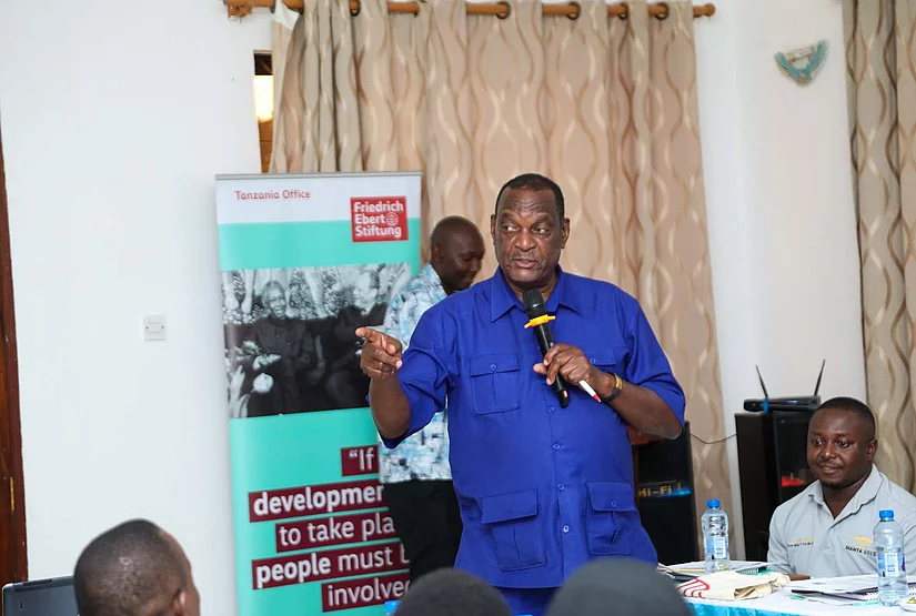 A man in a bright blue shirt speaks into a microphone and gestures at a small community meeting, with attendees listening; the mood is earnest and engaged.