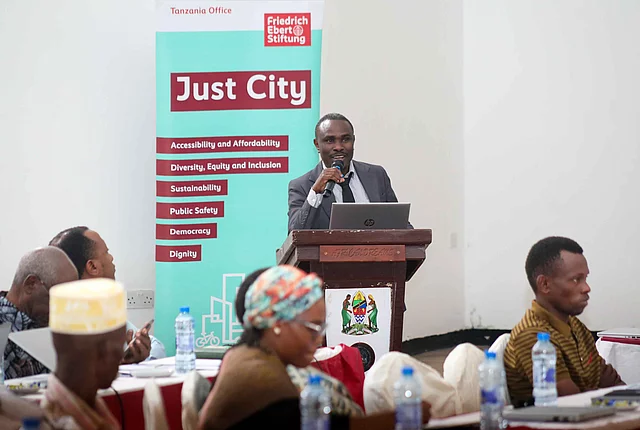 A man speaking at a conference, using a microphone while an audience listens attentively in the background.