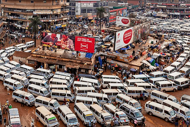 A busy urban transportation hub filled with tightly packed white minibuses used for public transit. People walk among the vehicles, indicating active movement and commerce. Surrounding the terminal are commercial buildings with signs and billboards, including advertisements for Coca-Cola and Airtel. The ground is unpaved and muddy, contributing to the gritty, vibrant atmosphere of the city.