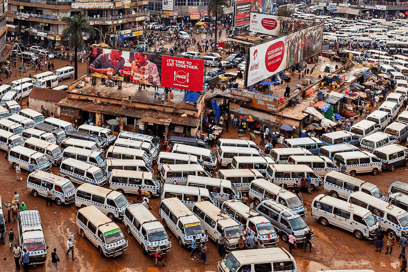 A busy urban transportation hub filled with tightly packed white minibuses used for public transit. People walk among the vehicles, indicating active movement and commerce. Surrounding the terminal are commercial buildings with signs and billboards, including advertisements for Coca-Cola and Airtel. The ground is unpaved and muddy, contributing to the gritty, vibrant atmosphere of the city.