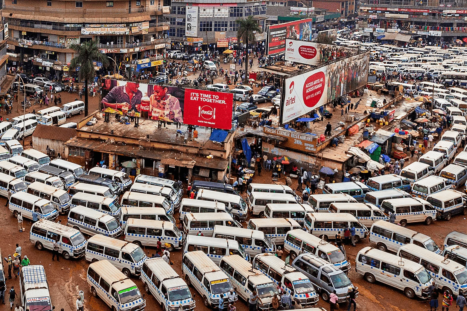 A busy urban transportation hub filled with tightly packed white minibuses used for public transit. People walk among the vehicles, indicating active movement and commerce. Surrounding the terminal are commercial buildings with signs and billboards, including advertisements for Coca-Cola and Airtel. The ground is unpaved and muddy, contributing to the gritty, vibrant atmosphere of the city.