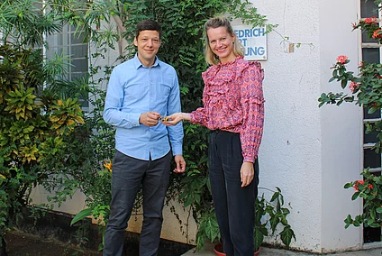 Two people stand outside near a building surrounded by plants and flowers. One person hands an object, possibly a card or paper, to the other.