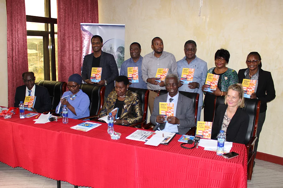 The authors of the publication together with the FES Team and the Guest of Honor pose together at a formal indoor event of a book launch. Six stand in the back while five sit at a table covered with a red cloth. Each person holds a copy of the book titled Building the Just City in Tanzania: Essays on Urban Housing, featuring a colorful red, yellow, and blue cover. The table displays water bottles, documents, and microphones, suggesting a press or panel setting. A partially visible banner in the background reinforces the organized and professional nature of the gathering.
