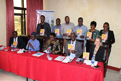 The authors of the publication together with the FES Team and the Guest of Honor pose together at a formal indoor event of a book launch. Six stand in the back while five sit at a table covered with a red cloth. Each person holds a copy of the book titled Building the Just City in Tanzania: Essays on Urban Housing, featuring a colorful red, yellow, and blue cover. The table displays water bottles, documents, and microphones, suggesting a press or panel setting. A partially visible banner in the background reinforces the organized and professional nature of the gathering.