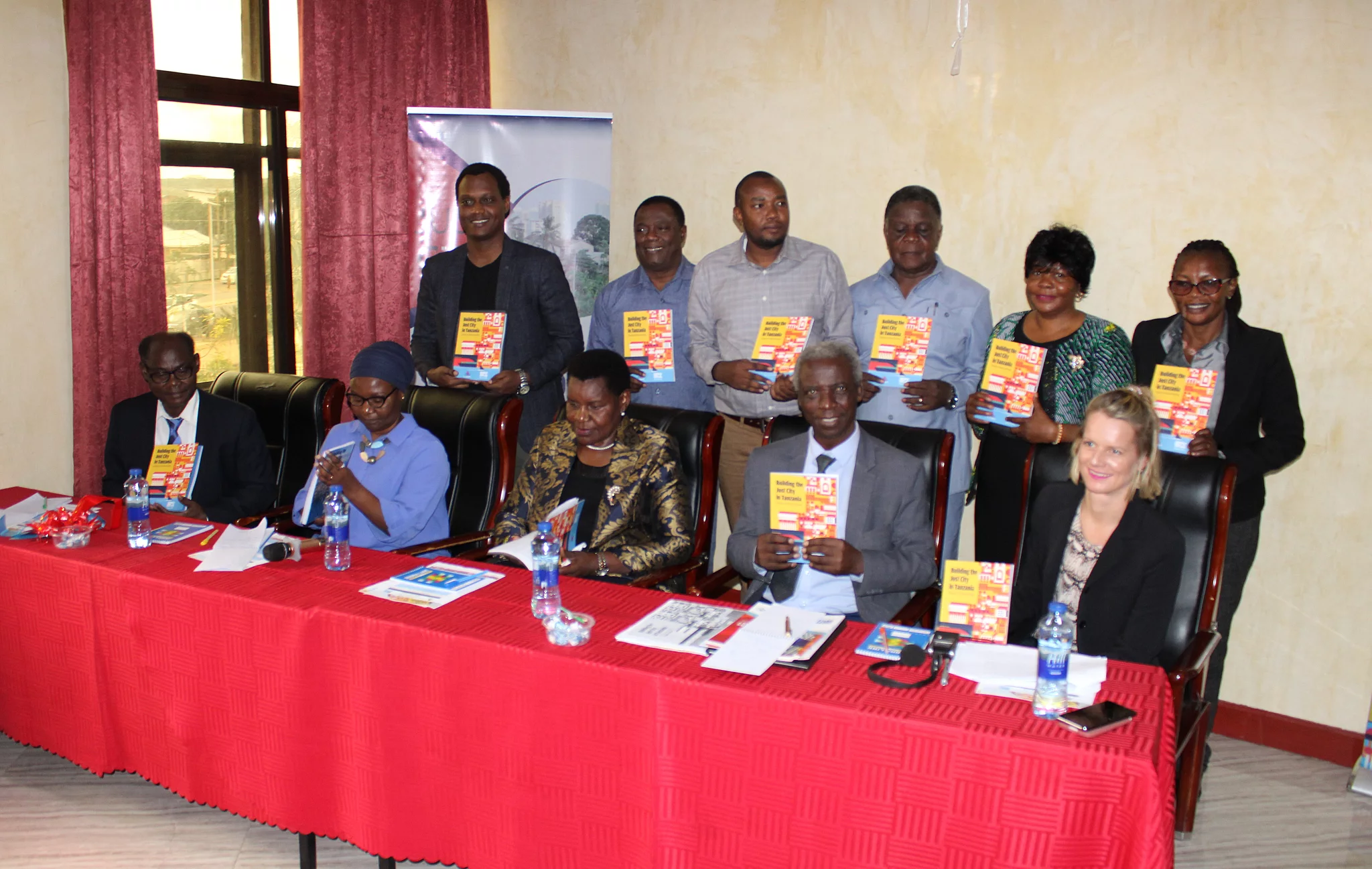 The authors of the publication together with the FES Team and the Guest of Honor pose together at a formal indoor event of a book launch. Six stand in the back while five sit at a table covered with a red cloth. Each person holds a copy of the book titled Building the Just City in Tanzania: Essays on Urban Housing, featuring a colorful red, yellow, and blue cover. The table displays water bottles, documents, and microphones, suggesting a press or panel setting. A partially visible banner in the background reinforces the organized and professional nature of the gathering.