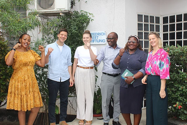Six people stand in a line outside a building, smiling and making a fist gesture with one hand. They are dressed in casual and semi-formal clothing. 