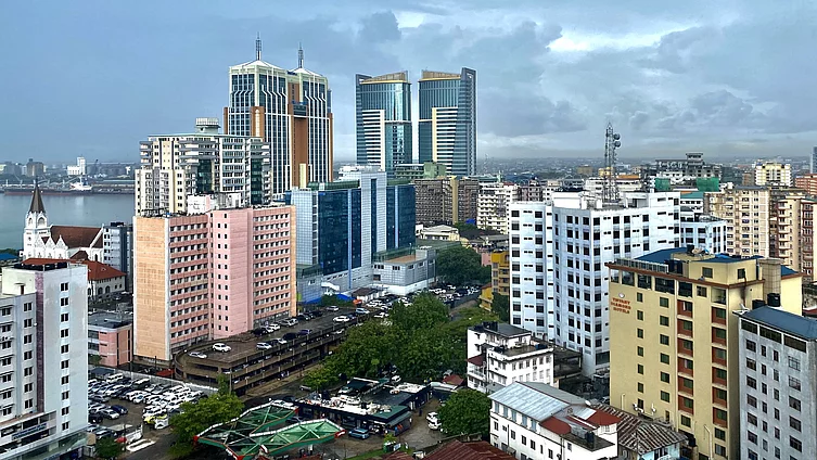 A coastal cityscape featuring a mix of modern high-rise buildings and older mid-rise structures. Prominent in the skyline are twin glass towers and a striking building with vertical orange and white stripes. In the foreground, a pink building and a white one labeled "TRINITY HOUSE" stand near a church with a red roof and pointed steeple. The area is densely built with parked vehicles and varied architecture, under an overcast sky suggesting recent or impending rain.