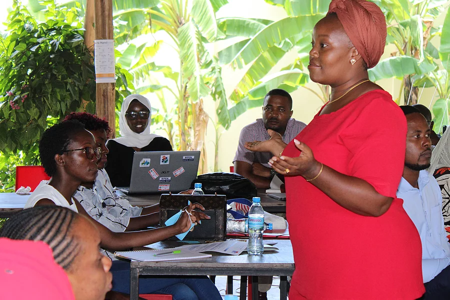 A group of people gathered in a semi-outdoor setting for a discussion or workshop. A woman in a red dress and headwrap stands and speaks to the seated participants, who have laptops and notebooks on the tables. The background features lush banana plants and wooden pillars, creating a tropical and informal learning environment.