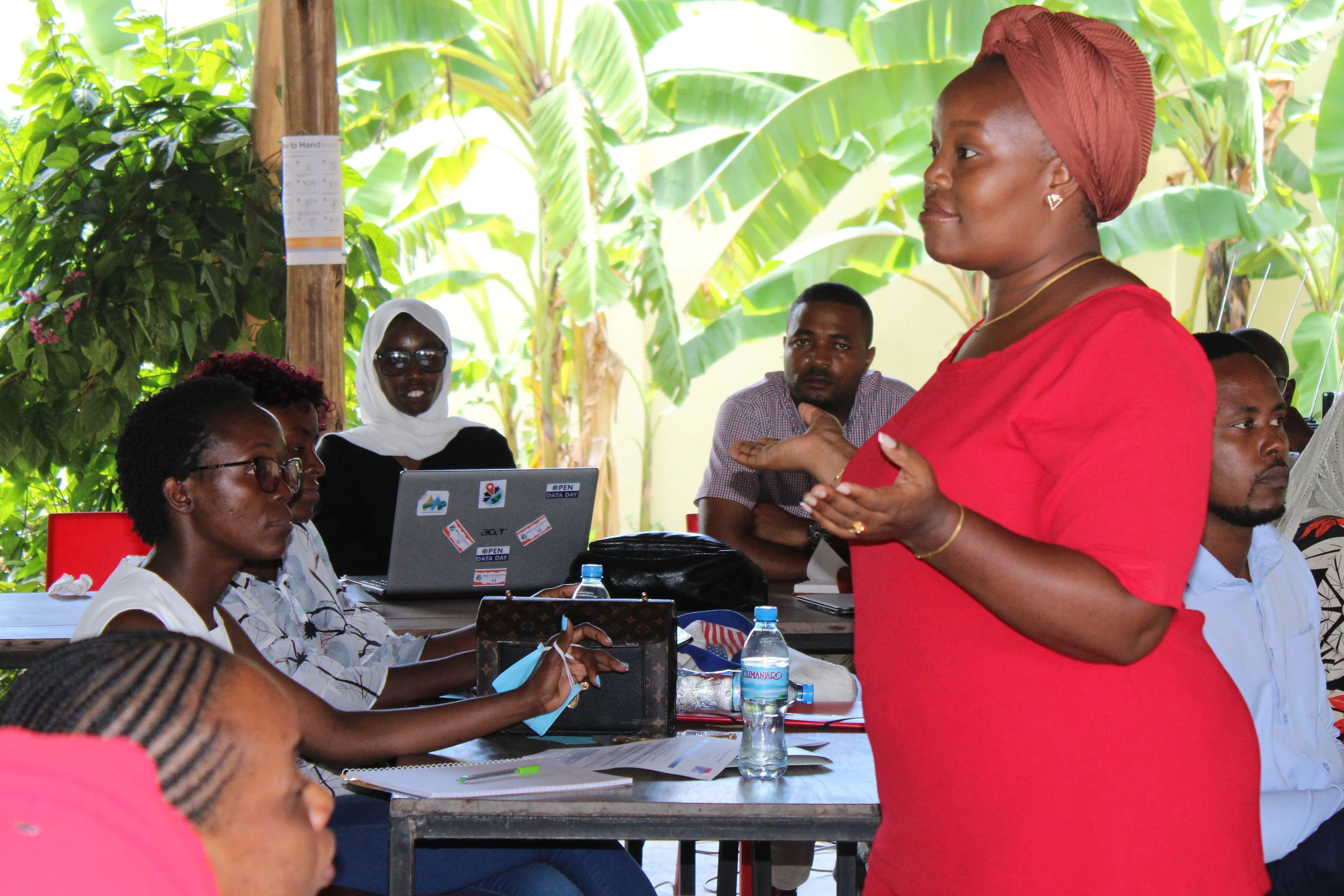 A group of people gathered in a semi-outdoor setting for a discussion or workshop. A woman in a red dress and headwrap stands and speaks to the seated participants, who have laptops and notebooks on the tables. The background features lush banana plants and wooden pillars, creating a tropical and informal learning environment.