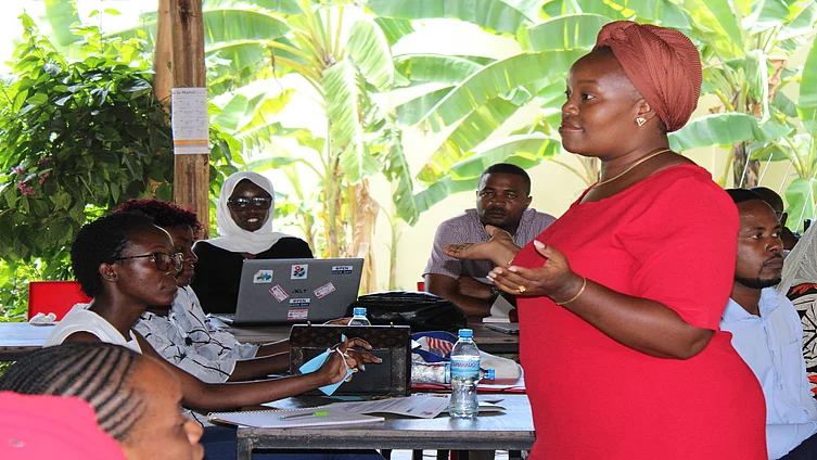 A group of people gathered in a semi-outdoor setting for a discussion or workshop. A woman in a red dress and headwrap stands and speaks to the seated participants, who have laptops and notebooks on the tables. The background features lush banana plants and wooden pillars, creating a tropical and informal learning environment.