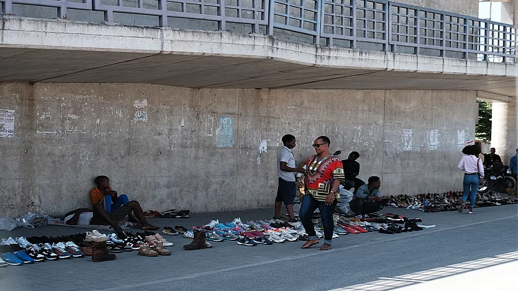 A street market scene beneath a concrete overpass where vendors display rows of shoes—sneakers, sandals, and dress shoes—directly on the pavement. People are seated near their merchandise or walking by, including one individual in a red patterned shirt and sunglasses. A large concrete wall with faded posters and a metal railing forms the background, highlighting informal urban commerce.