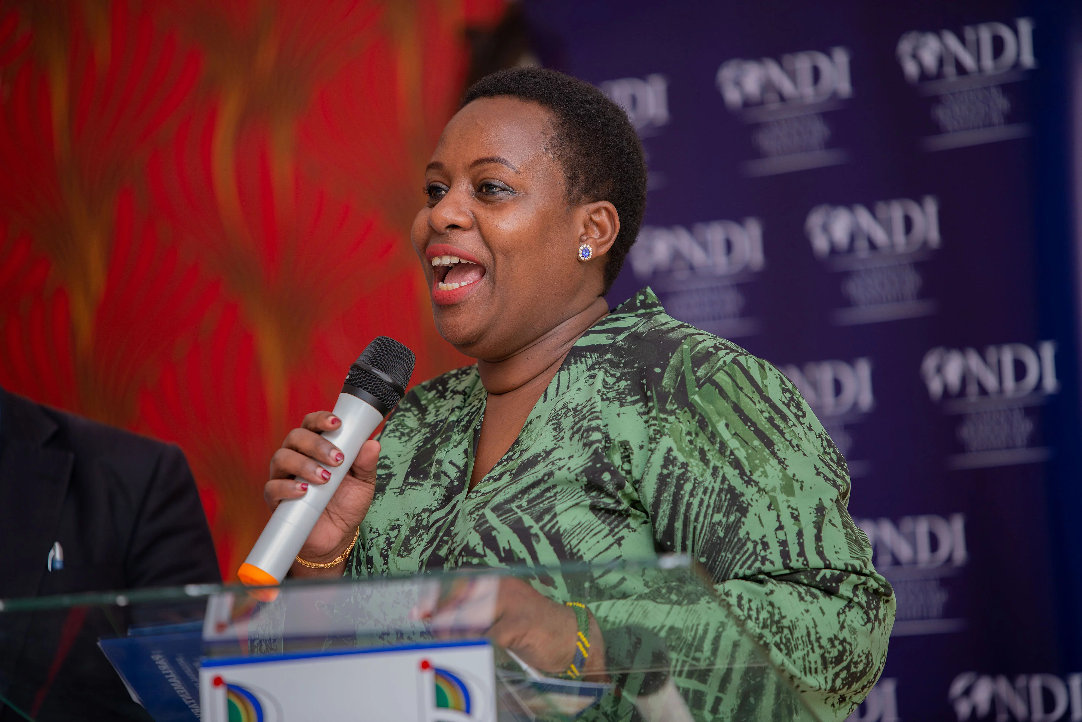 A person in a green patterned outfit speaks into a microphone at a podium during a formal event. The setting suggests a public speaking engagement such as a conference or seminar.
