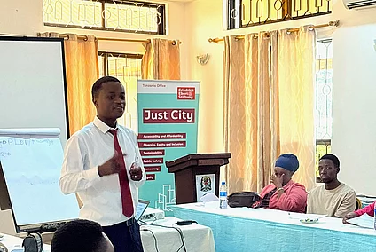 A conference or workshop setting with participants seated around tables covered in white and blue cloths. A man in a white shirt and red tie stands and speaks in front of the group, possibly presenting or leading a discussion. Behind him are a podium, a flip chart, a projector screen, and a banner that reads "Boot Camp." The room has a formal and educational atmosphere focused on professional development or training.