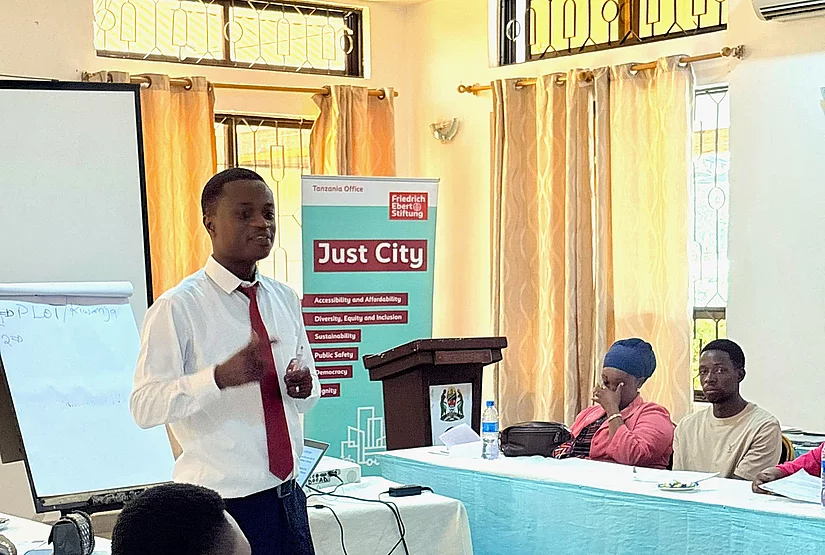 A conference or workshop setting with participants seated around tables covered in white and blue cloths. A man in a white shirt and red tie stands and speaks in front of the group, possibly presenting or leading a discussion. Behind him are a podium, a flip chart, a projector screen, and a banner that reads "Boot Camp." The room has a formal and educational atmosphere focused on professional development or training.