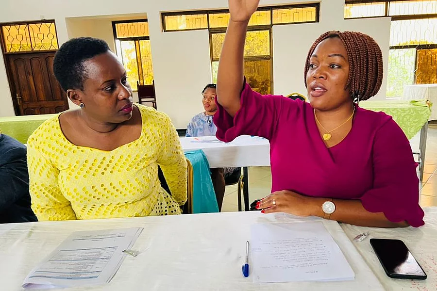 Two individuals seated at a table in a meeting room setting. One, dressed in yellow, looks attentively at the other, who wears magenta and has one hand raised, possibly making a point or asking a question. Papers, a pen, and a smartphone lie on the table between them. In the background, other participants are seated and appear engaged, suggesting an active, collaborative discussion environment.
