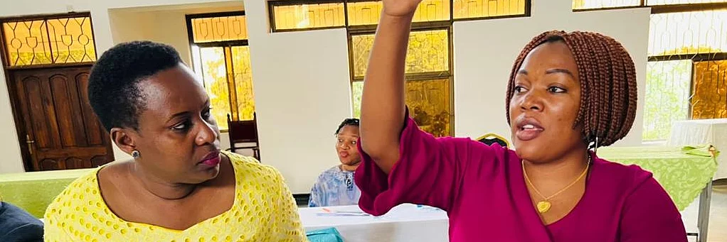 Two individuals seated at a table in a meeting room setting. One, dressed in yellow, looks attentively at the other, who wears magenta and has one hand raised, possibly making a point or asking a question. Papers, a pen, and a smartphone lie on the table between them. In the background, other participants are seated and appear engaged, suggesting an active, collaborative discussion environment.