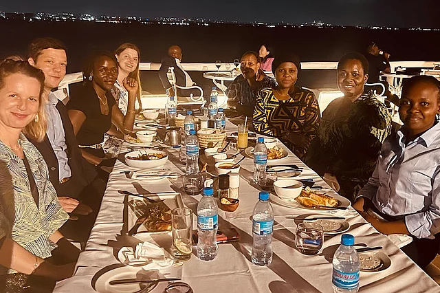 A group of people sit around a long outdoor dining table at night, set with plates of food, glasses, and bottles of water. Behind them, a body of water reflects distant city lights, creating a scenic waterfront backdrop. The atmosphere suggests a formal or semi-formal dinner gathering with a communal and celebratory feel.
