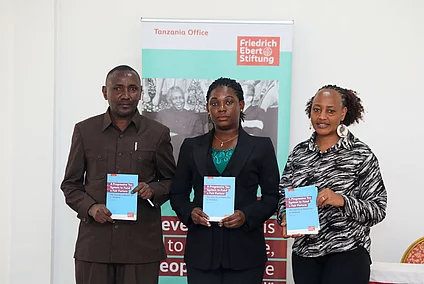 Three individuals stand holding books in front of a banner for the Friedrich Ebert Stiftung Tanzania Office, conveying professionalism and focus.