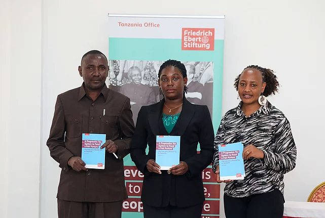 Three individuals stand holding books in front of a banner for the Friedrich Ebert Stiftung Tanzania Office, conveying professionalism and focus.