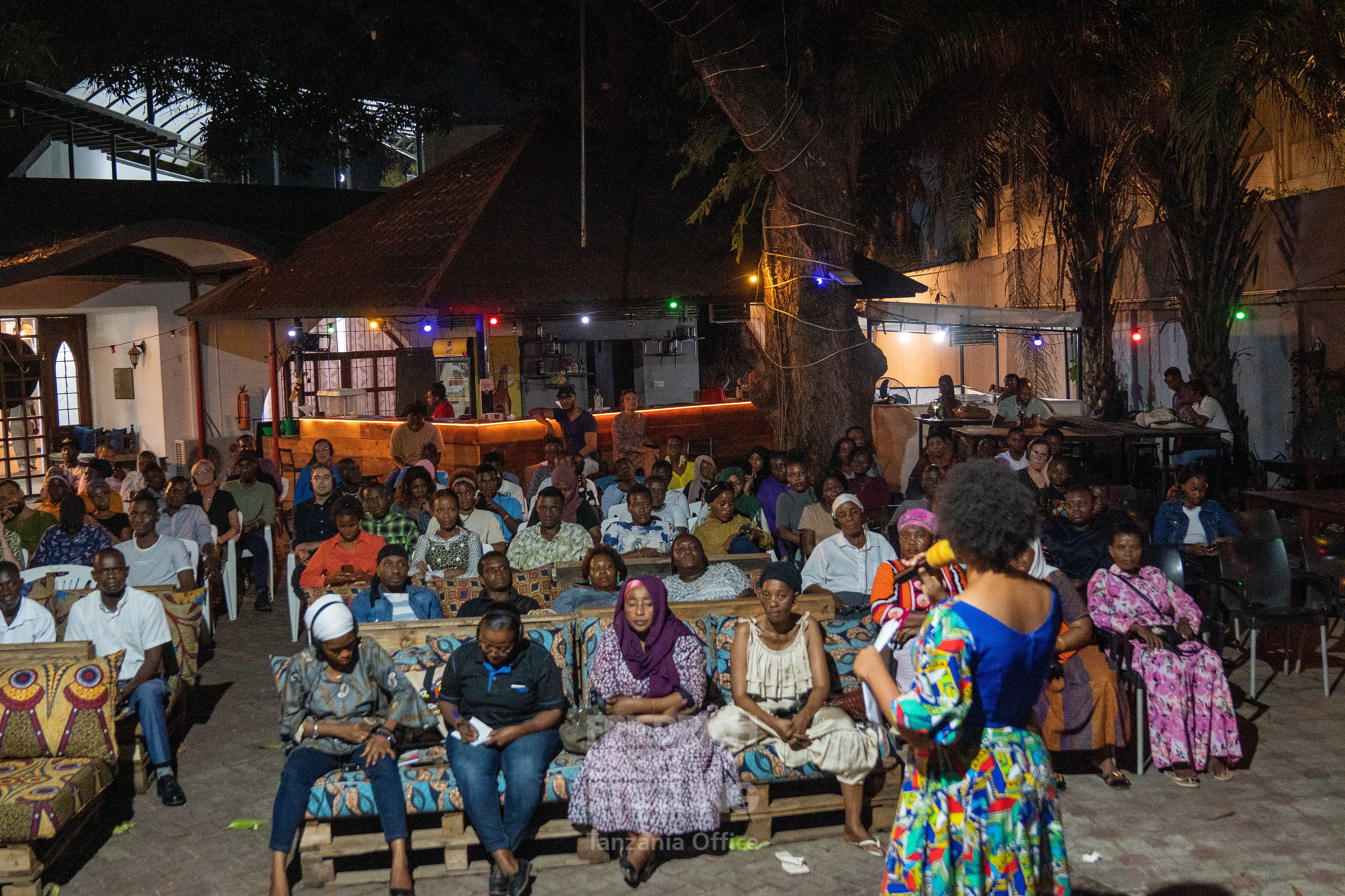 A nighttime outdoor event with rows of seated attendees facing a speaker in a colorful outfit. The setting is adorned with string lights among trees and illuminated buildings in the background, creating a festive and welcoming atmosphere. The diverse attire and attentive audience suggest a cultural or community gathering.