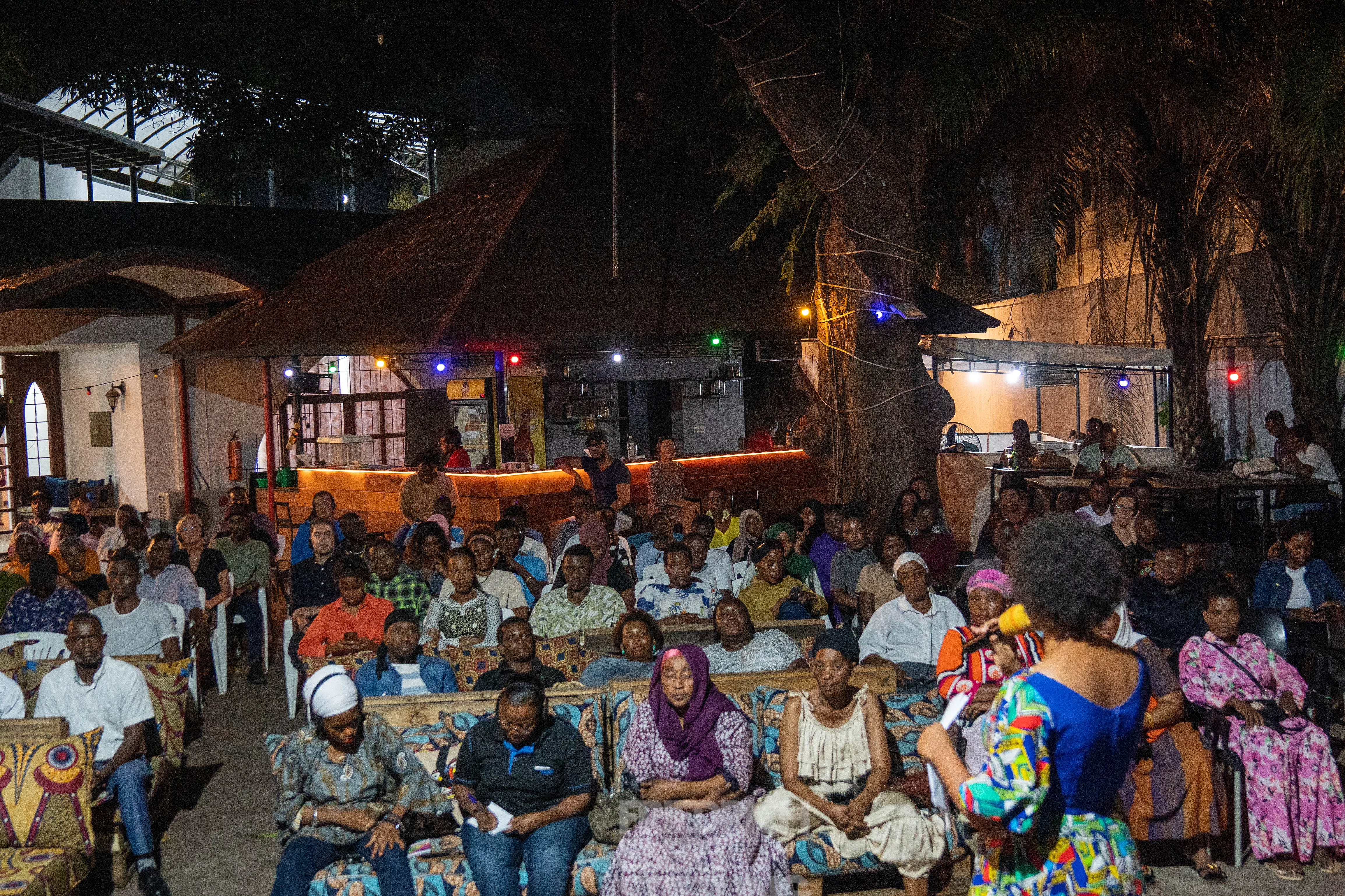 A nighttime outdoor event with rows of seated attendees facing a speaker in a colorful outfit. The setting is adorned with string lights among trees and illuminated buildings in the background, creating a festive and welcoming atmosphere. The diverse attire and attentive audience suggest a cultural or community gathering.