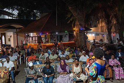 A nighttime outdoor event with rows of seated attendees facing a speaker in a colorful outfit. The setting is adorned with string lights among trees and illuminated buildings in the background, creating a festive and welcoming atmosphere. The diverse attire and attentive audience suggest a cultural or community gathering.