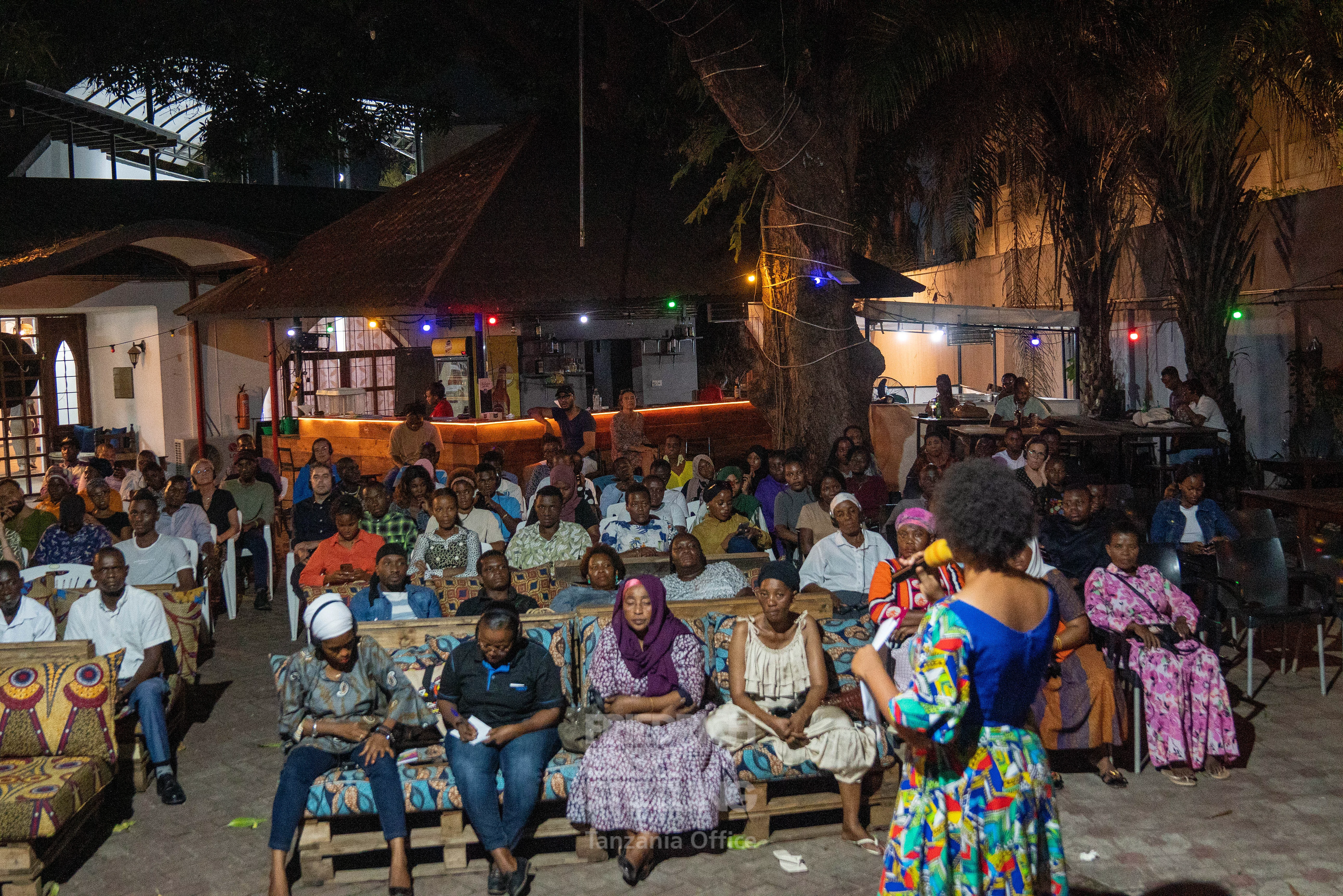 A nighttime outdoor event with rows of seated attendees facing a speaker in a colorful outfit. The setting is adorned with string lights among trees and illuminated buildings in the background, creating a festive and welcoming atmosphere. The diverse attire and attentive audience suggest a cultural or community gathering.