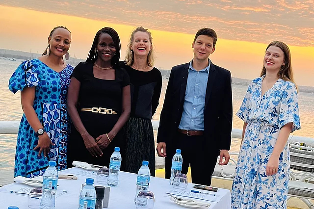 Five people stand behind a table set for a meal outdoors at sunset by the ocean. The sky glows with orange and pink hues. The table, covered with a white cloth, holds water bottles, glasses, and plates. The group is dressed in semi-formal attire, suggesting a celebratory or special occasion.