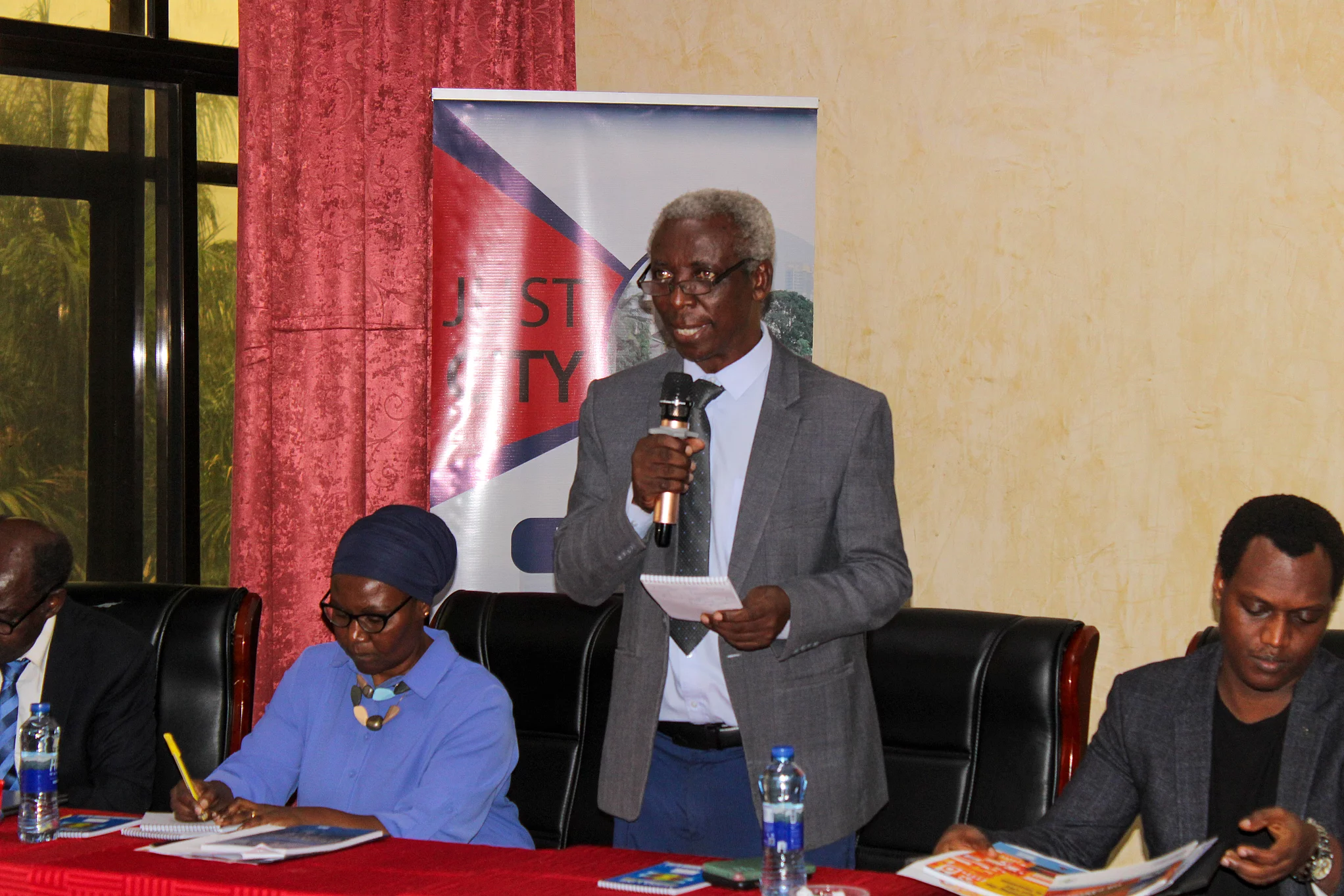 A group of individuals seated at a table during a formal meeting or conference. One person stands at the center, speaking while holding a piece of paper. The background features a banner with partially visible text reading "Just City" suggesting the event relates to Urbanization initiatives. The participants are dressed in formal attire, with water bottles and documents on the table, indicating a professional and organized setting.