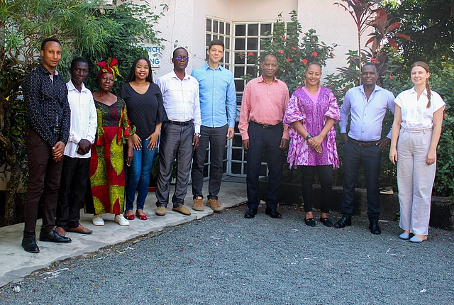 A group of ten FES Staff in Tanzania stands in a line outdoors in front of a building entrance, dressed in a mix of formal and traditional attire. Behind them are green plants and flowers, with part of a sign reading "Friedrich-Ebert-Stiftung" visible on the wall. The setting suggests a professional or cultural gathering in a welcoming, community-oriented environment.