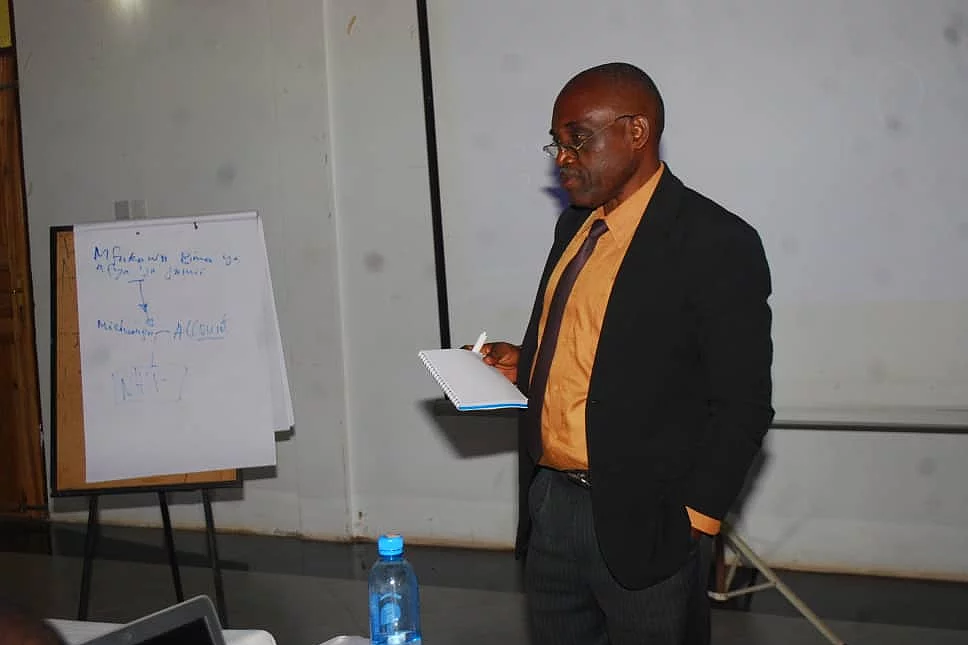 A man stands in a seminar room, holding a piece of paper as he presents. He wears a black suit with an orange shirt. To his left is a flip chart with handwritten text. In front of him is a table with a blue water bottle and electronic equipment. A projector screen is visible in the background, reinforcing the educational setting focused on research methods.