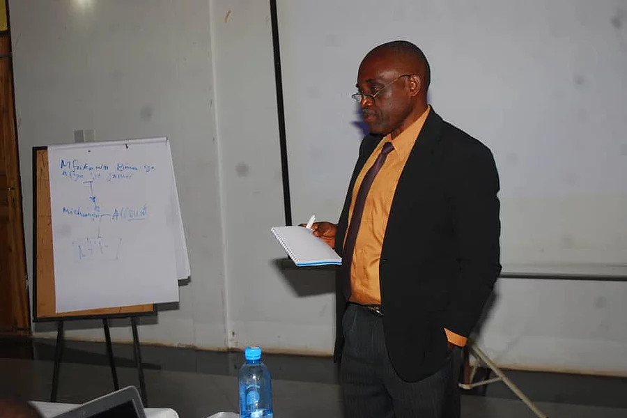 A man stands in a seminar room, holding a piece of paper as he presents. He wears a black suit with an orange shirt. To his left is a flip chart with handwritten text. In front of him is a table with a blue water bottle and electronic equipment. A projector screen is visible in the background, reinforcing the educational setting focused on research methods.