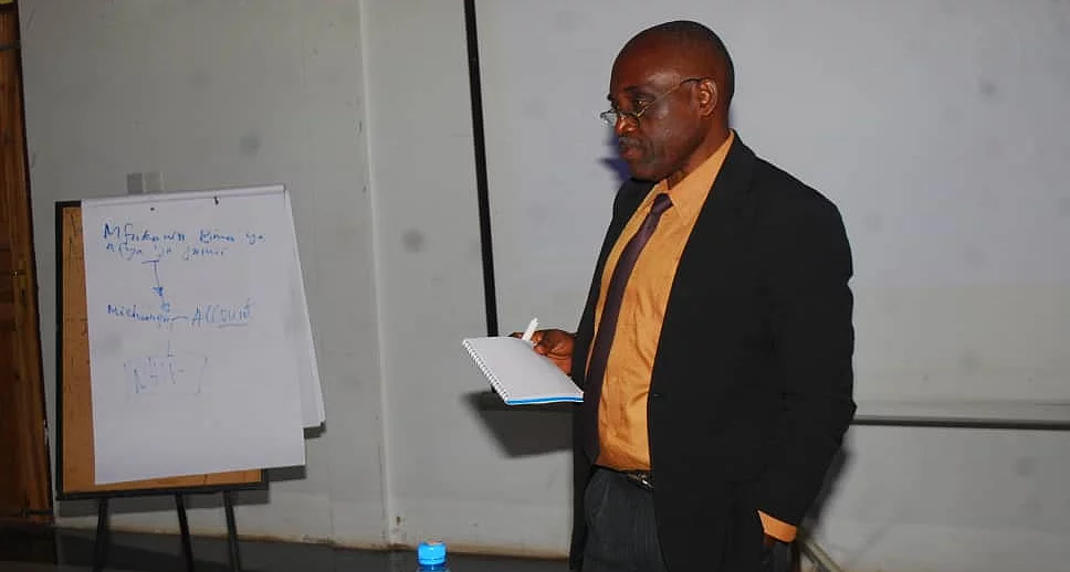 A man stands in a seminar room, holding a piece of paper as he presents. He wears a black suit with an orange shirt. To his left is a flip chart with handwritten text. In front of him is a table with a blue water bottle and electronic equipment. A projector screen is visible in the background, reinforcing the educational setting focused on research methods.