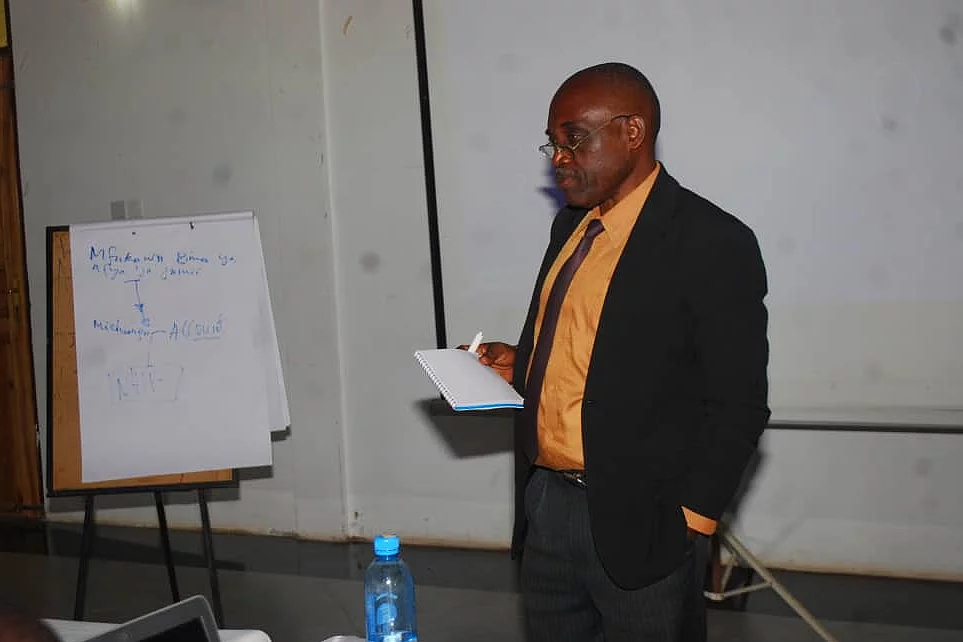 A man stands in a seminar room, holding a piece of paper as he presents. He wears a black suit with an orange shirt. To his left is a flip chart with handwritten text. In front of him is a table with a blue water bottle and electronic equipment. A projector screen is visible in the background, reinforcing the educational setting focused on research methods.